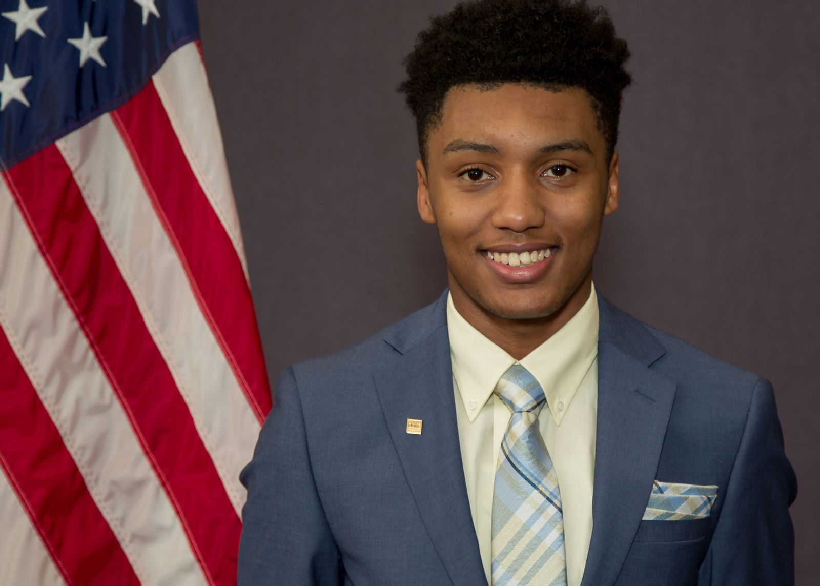 African American college student Rah'Mere Williams, dressed in a navy blue suit with a white collared shirt and light blue striped tie stands in front of and to the right of an American Flag against a grey background.