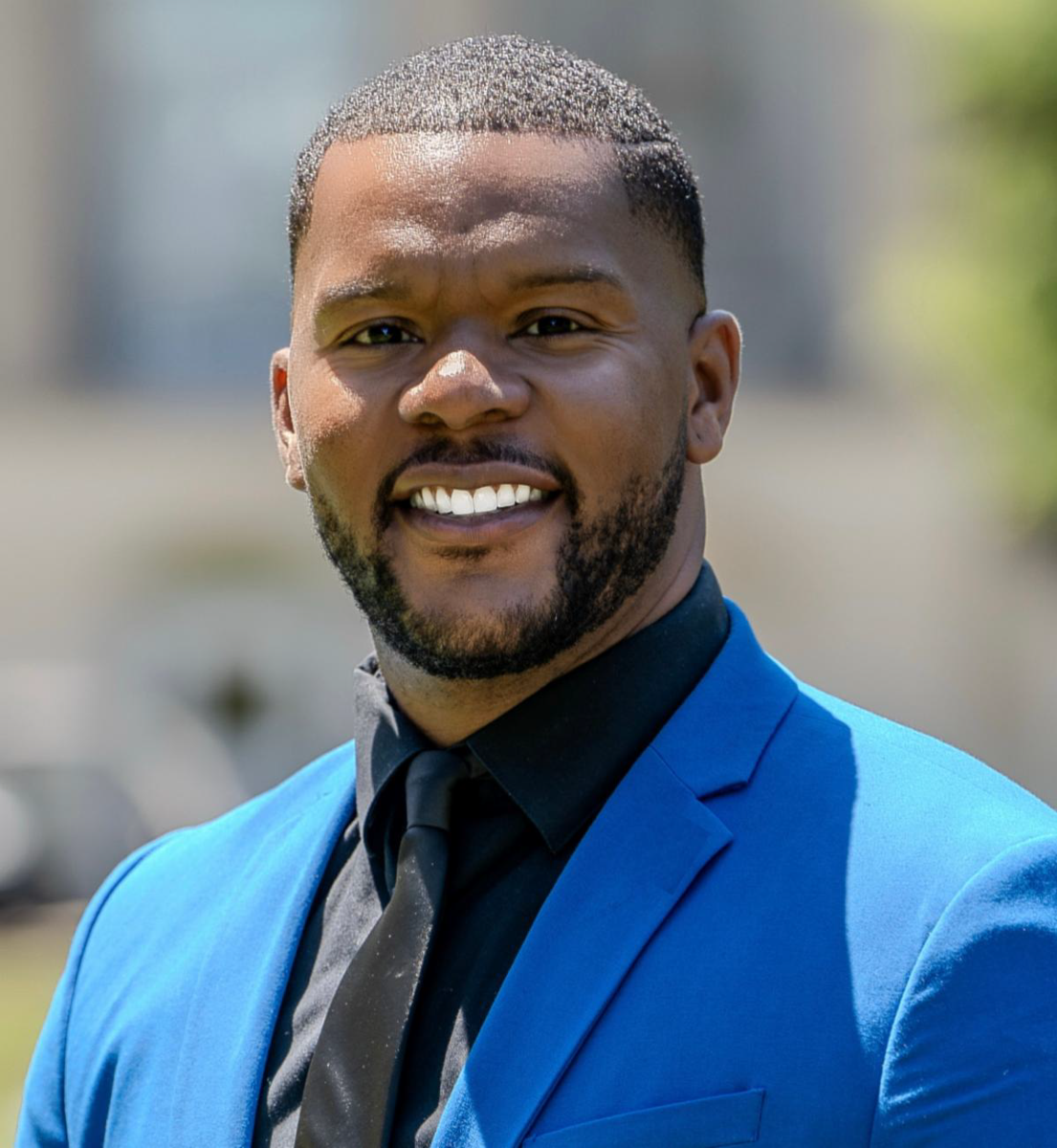 Dr. Kenvoris Blair, an African American Male, smiles at the camera wearing a blue suit with a black collared shirt and tie.