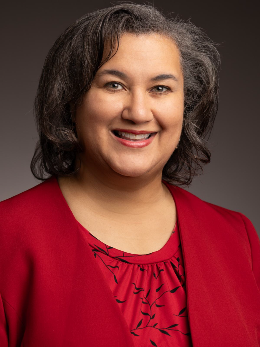 Dr. Melissa Frank-Alston, a Caucasian female with shoulder length brown and gray hair, smiles at the camera wearing a red dress jack and a red blouse with a black ivy pattern.