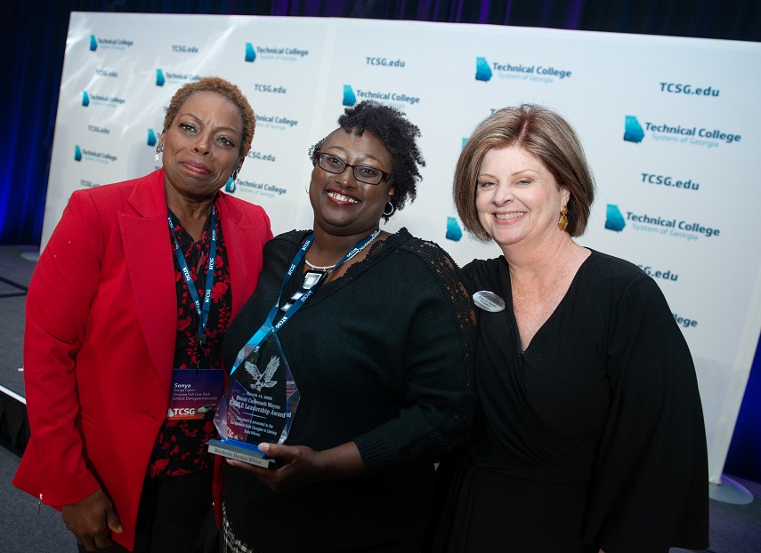 Smiling African American female with short brown hair wearing a red jacket, red and black dress, wearing a black and white lanyard around her neck, standing next to an African American female wearing black glasses, black cardigan and black and white dress, wearing a lanyard, holding a glass award, standing next to a Caucasian female with brown hair wearing a black dress, and a name badge posing for a photo against a white backdrop with the technical college system of georgia logo and tcsg dot edu in repeating patters in a teal color