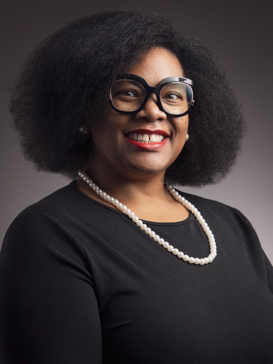 Ms. Sheffie Robinson, an African American female, wears a black dress shirt with pearls and black rimmed glasses while smiling at the camera.